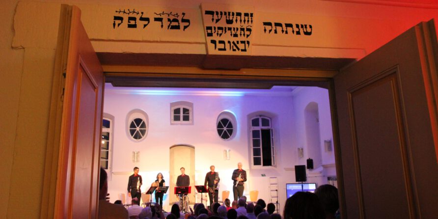 Eine Gruppe sieht fünf Musikern in einem hellen Saal durch Türen mit hebräischer Schrift zu.