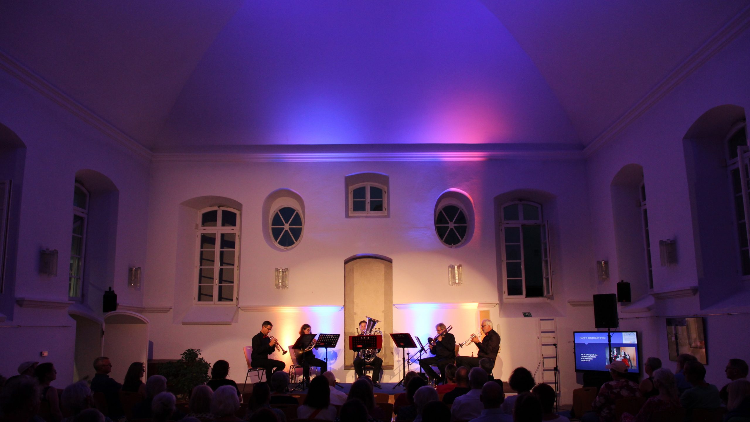 Kammermusik-Ensemble spielt vor Publikum auf heller Bühne mit hohen Decken, violettes Licht, Screen rechts.