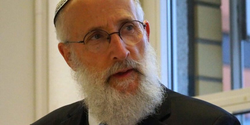 Rabbiner Jechiel Brukner, ein älterer Mann mit grauem Bart, Brille und Kippa, trägt Anzug und Krawatte. Er sitzt in einem hellen Raum vor einem Fenster und blickt nachdenklich zur Seite.