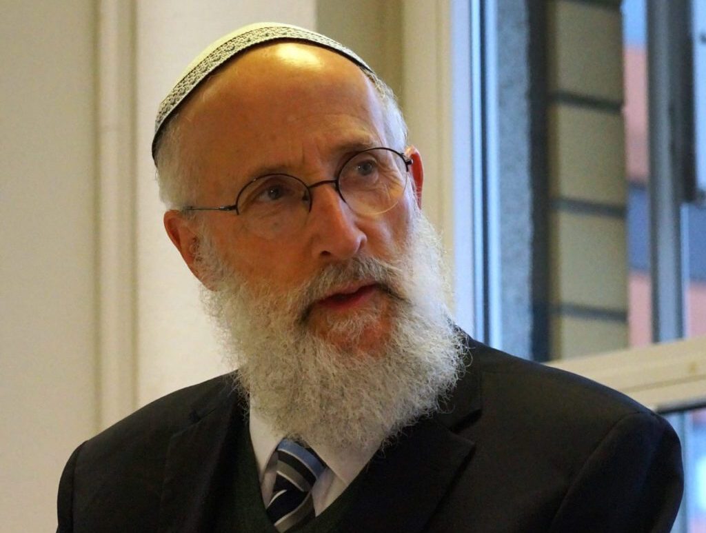 Rabbiner Jechiel Brukner, ein älterer Mann mit grauem Bart, Brille und Kippa, trägt Anzug und Krawatte. Er sitzt in einem hellen Raum vor einem Fenster und blickt nachdenklich zur Seite.