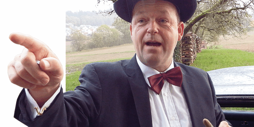 Ein Mann in festlicher Kleidung mit schwarzem Anzug, weißem Hemd, roter Fliege und schwarzem Hut steht im Freien. Er zeigt mit der rechten Hand nach vorne und hält in der linken Hand einen kleinen Gegenstand. Im Hintergrund sind ein Auto, grüne Wiesen und Bäume zu sehen.