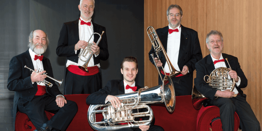 Fünf Männer in festlicher Kleidung mit roten Fliegen und schwarzen Anzügen sitzen und stehen um ein rotes Sofa. Jeder hält ein Blechblasinstrument, darunter Trompete, Horn, Posaune und Tuba. Sie lächeln in die Kamera. Der Hintergrund besteht aus einer Holz- und einer dunklen Wand.