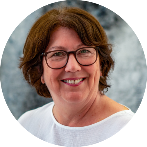 Hildegard Gooss mit brünetten Haaren und Brille lächelt freundlich vor blau-grauem Hintergrund.