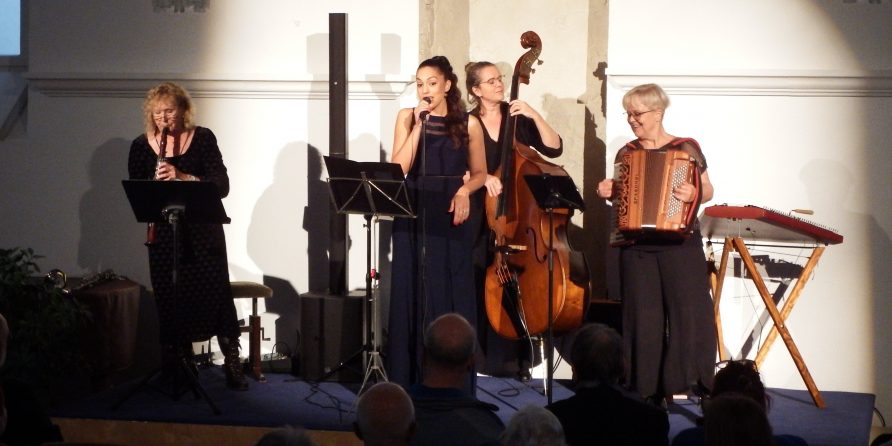 Vier Frauen musizieren auf der Bühne, das Publikum schaut in einem hellen Raum zu.