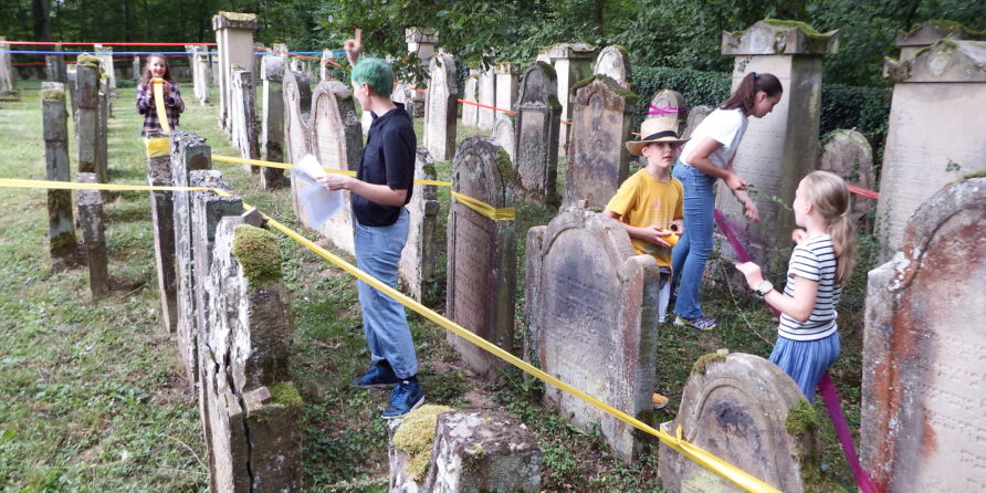 Kinder und Erwachsene erkunden mit Papieren einen alten Friedhof, gelbes Band sperrt Bereiche ab.