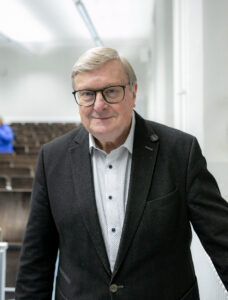 Professor Dr. Karl-Josef Kuschel mit Brille und grauen Haaren steht im leeren Hörsaal vor leeren Sitzreihen.