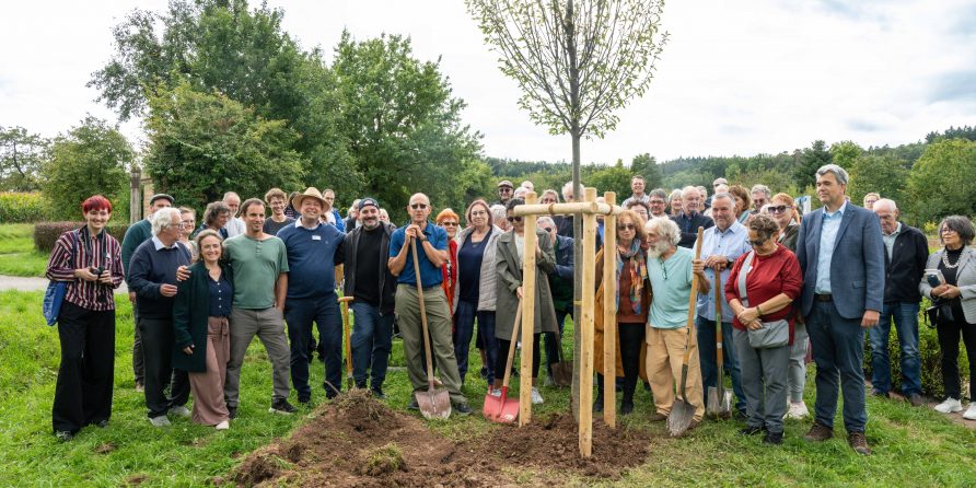 Eine große Gruppe steht draußen um einen frisch gepflanzten Baum mit Schaufeln im Gras.