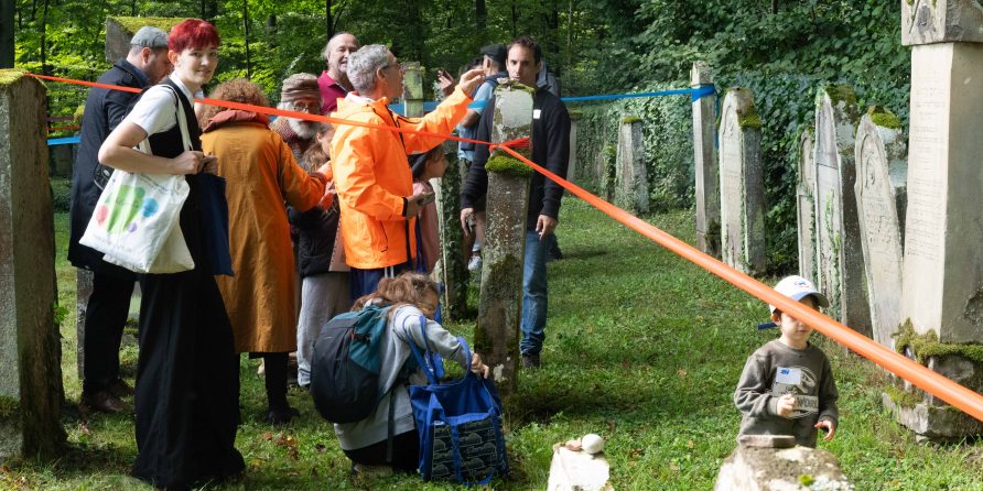 Menschen gehen zwischen alten Grabsteinen mit bunten Bändern, ein Kind mit grüner Mütze vorne.