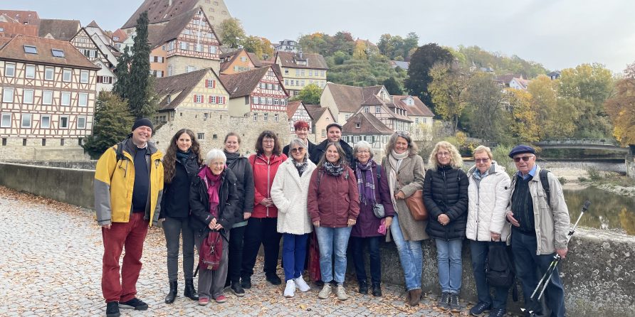 Eine Gruppe posiert lächelnd auf einem Steinweg vor Fachwerkhäusern im Herbst, Fluss im Hintergrund.