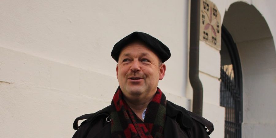 Ein Mann mit schwarzem Barett, dunklem Mantel und Schal steht vor weißer Wand und lächelt leicht.