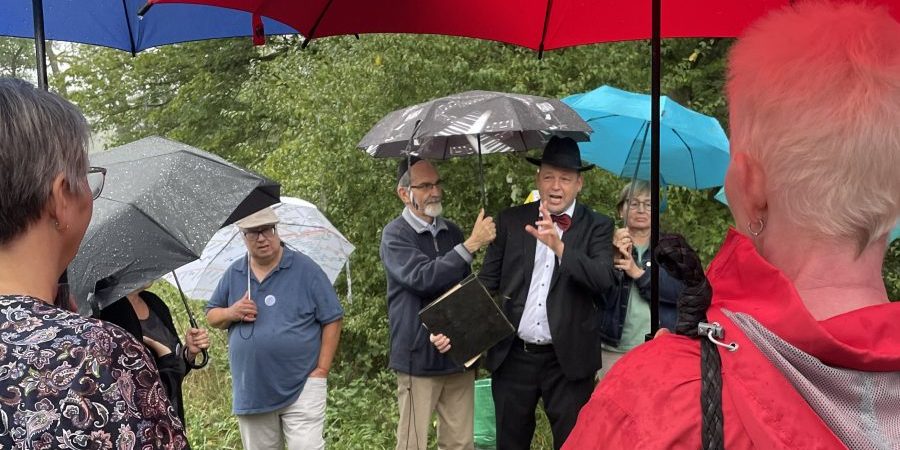 Menschen mit Regenschirmen stehen draußen im Regen; ein Mann im Anzug spricht gestikulierend.
