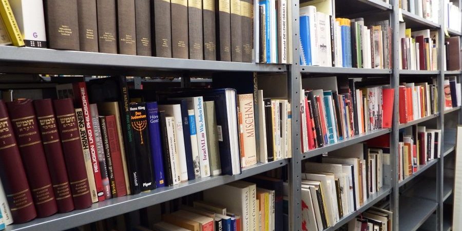 Bücherregale voller Bücher, einige stehen, andere lehnen; teils hebräische Titel in hellem Raum.