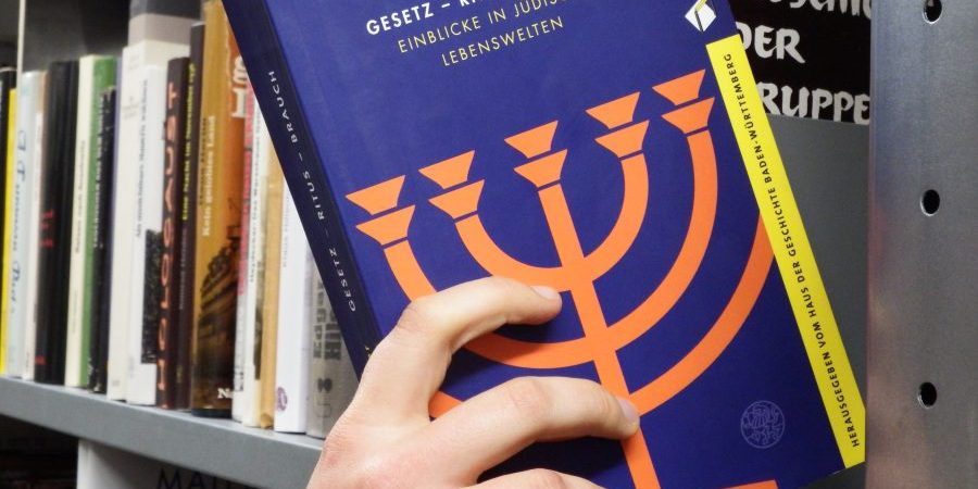 Eine Hand hält ein blaues Buch mit oranger Menora vor einem Bücherregal, deutscher Titel.
