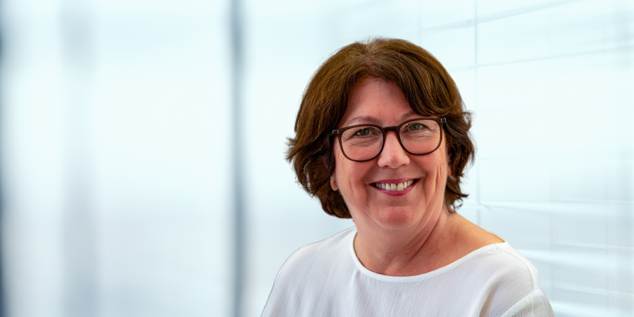 Hildegard Gooss mit brünetten Haaren und Brille lächelt freundlich vor unscharfem, hellem Hintergrund.